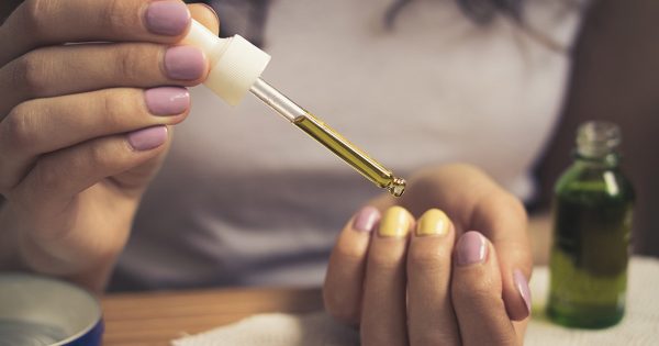 Female hands take care of cuticles with oil. Girl in white t-shirt sitting at the table and takes care of her cuticles. She has a bright manicure. On the table lay manicure equipment. Vintage filter